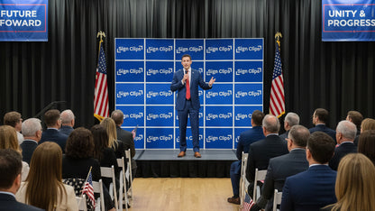 Candidate standing on a stage with a Sign Clip Wall made of yard signs behind him, addressing an audience.